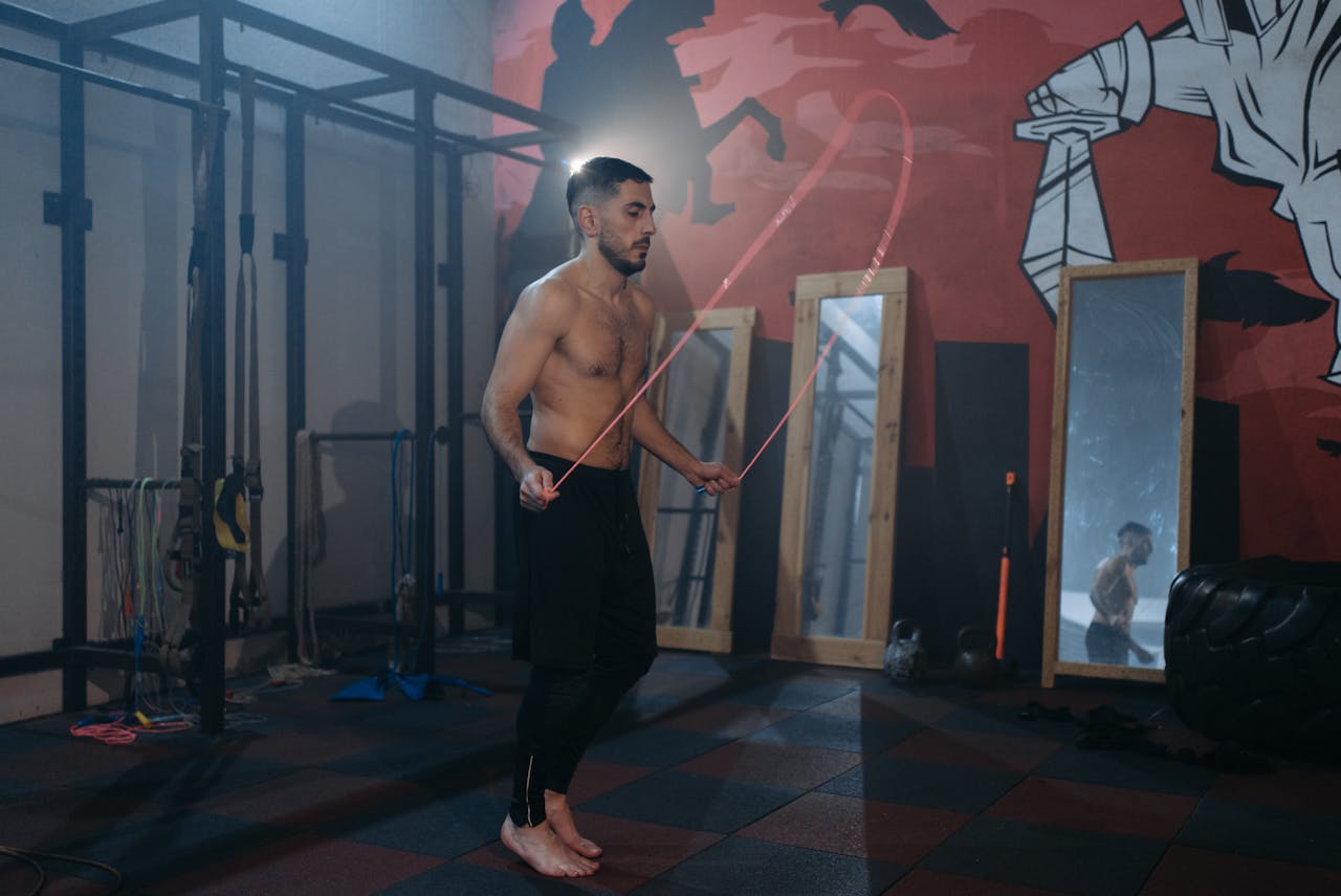 A muscular man performs jump rope exercise in a gym surrounded by mirrors.